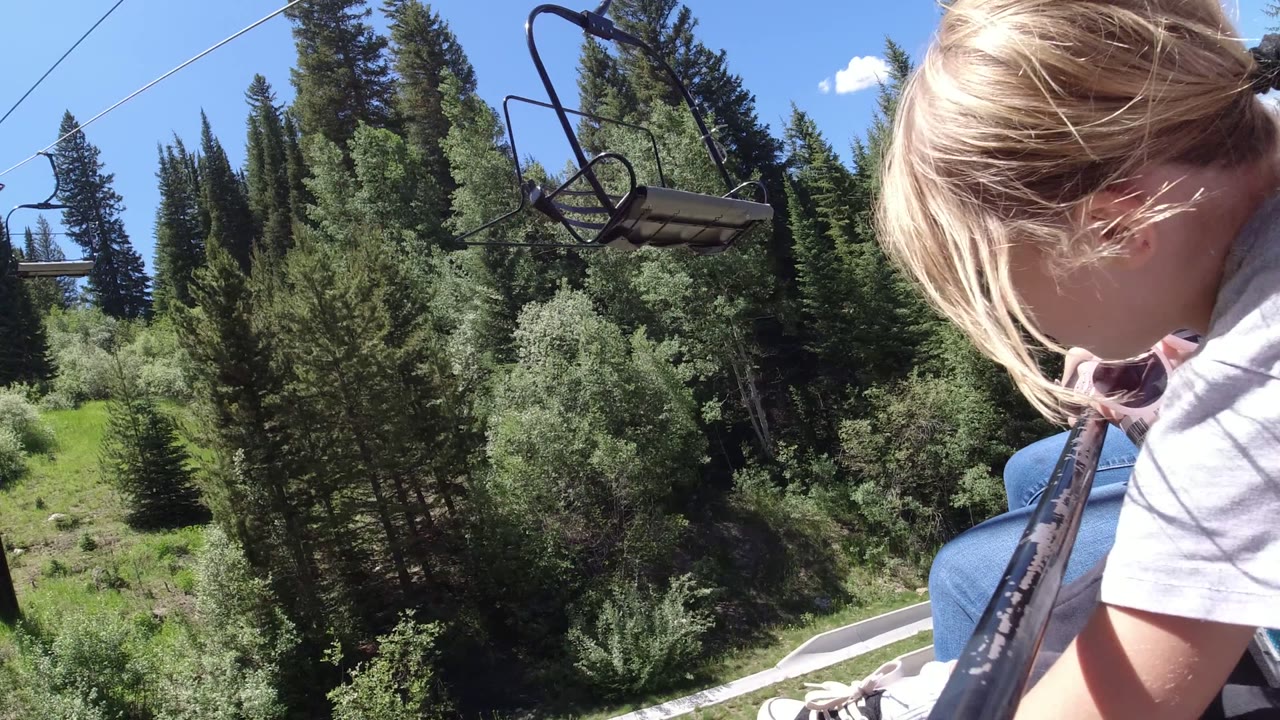 A blond child sits close to the camera on an open lift in Winter Park, Colorado, with adults nearby, a black rail in the foreground, forest below, and blue sky above.