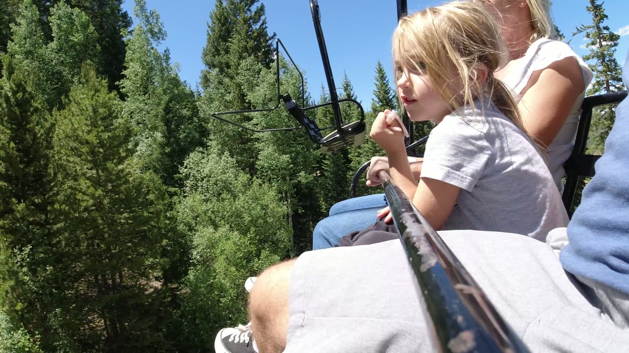 The child turns slightly on the ropeway seat in Winter Park, Colorado, while adults remain partly cropped and dense green trees fill the background.
