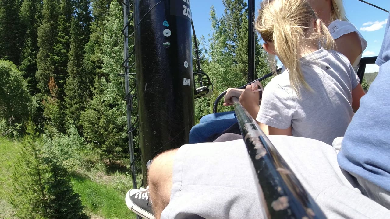 The ride seat passes close to a black vertical pole in Winter Park, Colorado, while the family remains partly visible beside the foreground safety bar and leafy hillside.