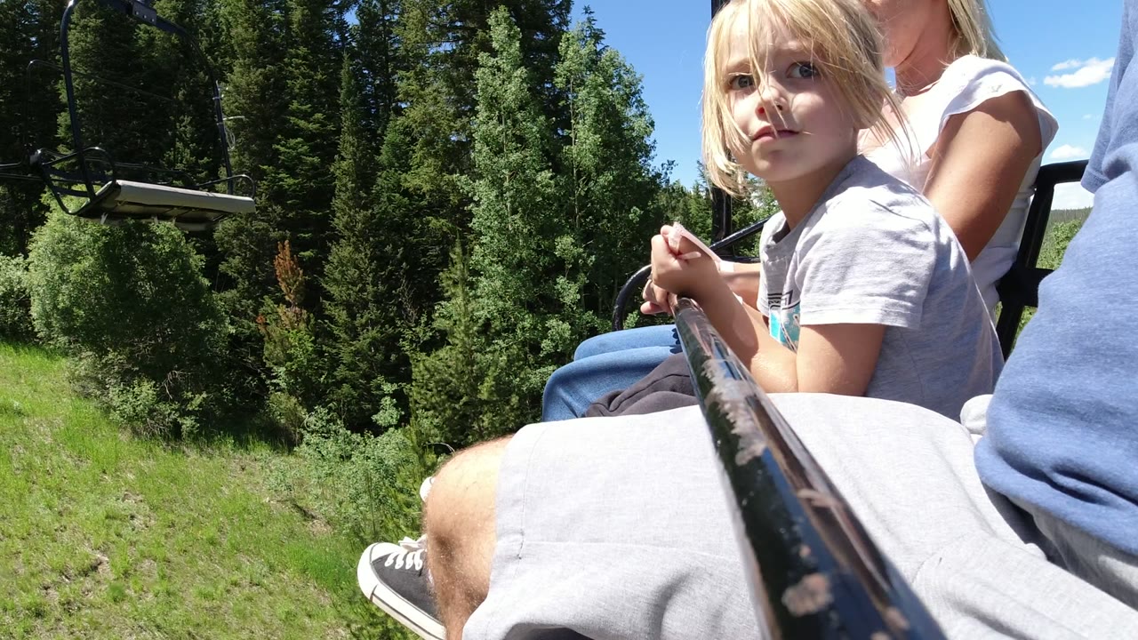 Child and adults seated on an open ropeway seat above a grassy forest slope.