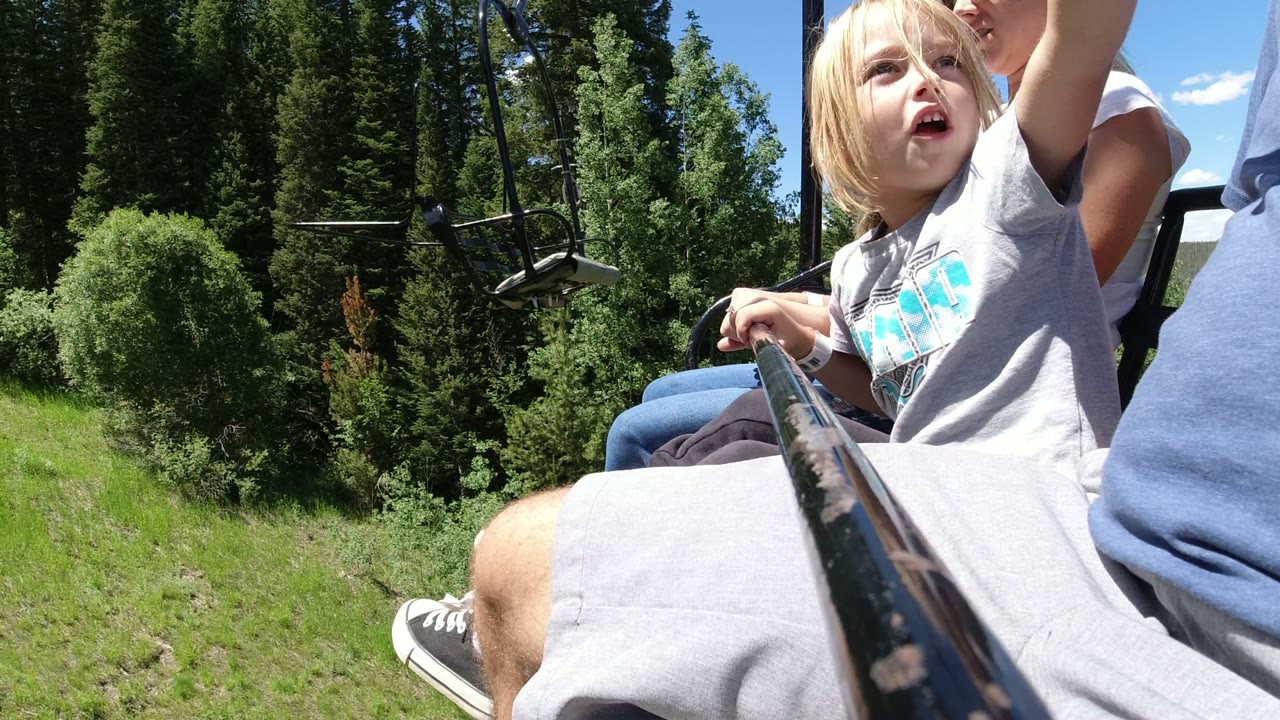 At 0:01, the child lifts an arm while seated between adults on the Winter Park ropeway, with the foreground bar pointing toward a green slope and tall trees.