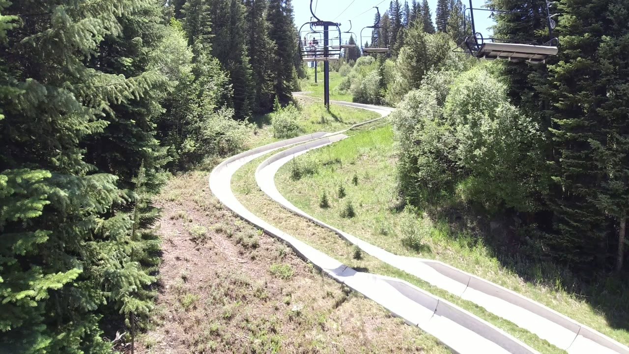 Elevated view of a pale alpine slide track curving through green hillside and evergreen forest in Winter Park, Colorado, with chairlift structures in the distance and no visible people.