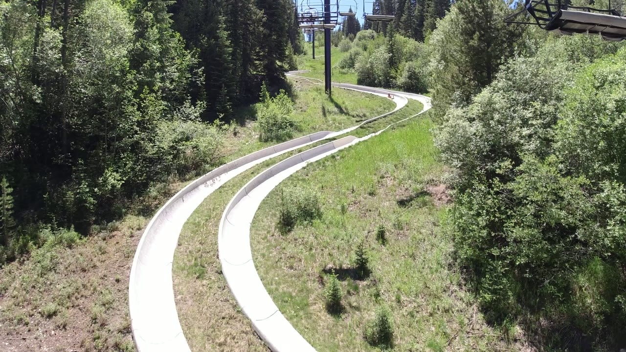 The empty pale slide tracks bend through bright grass and thick forest in Winter Park, Colorado, with no visible people in this moment.