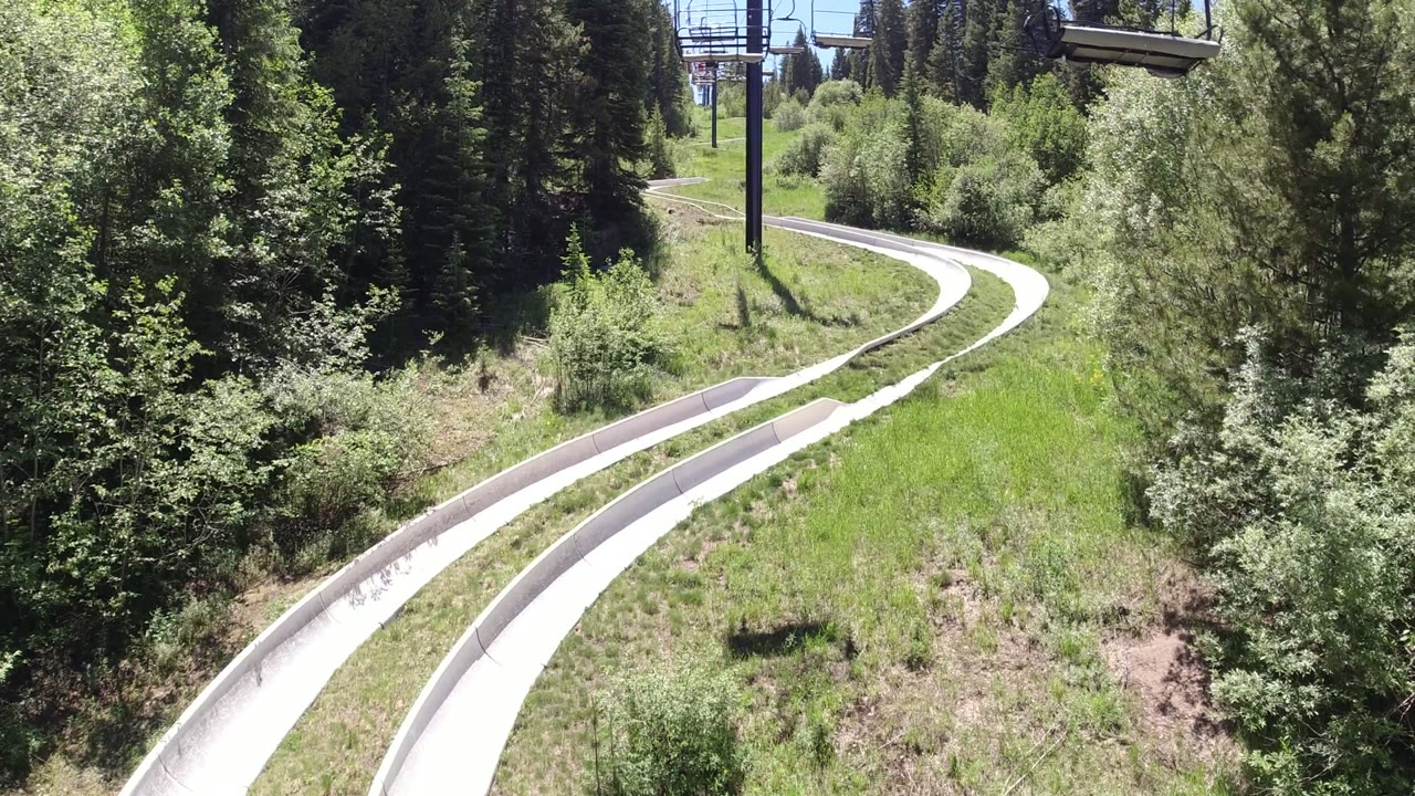 At 0:00 in Winter Park, Colorado, pale curved track lanes run through green grass and dense trees beneath overhead cableway hardware, with no visible people.