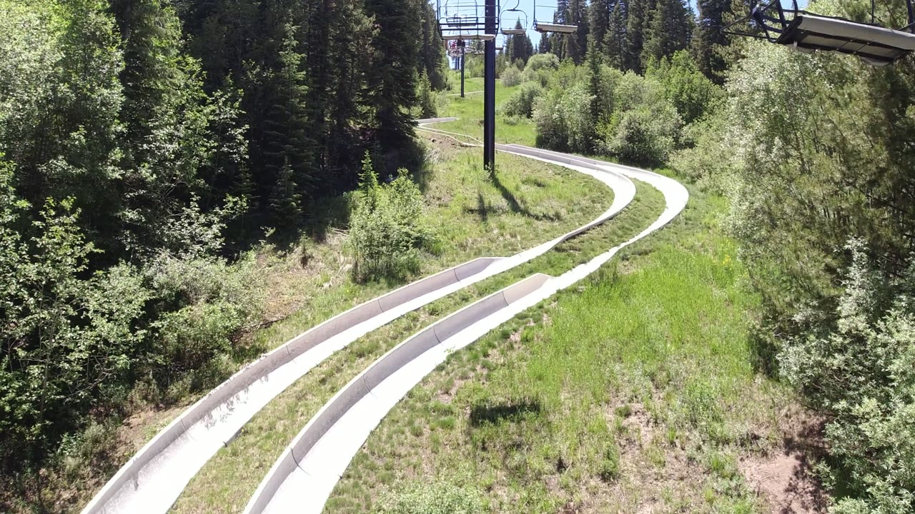 At 0:01 in Winter Park, Colorado, the parallel pale lanes curve deeper into a green corridor of grass, shrubs, and evergreen trees, with no visible people.