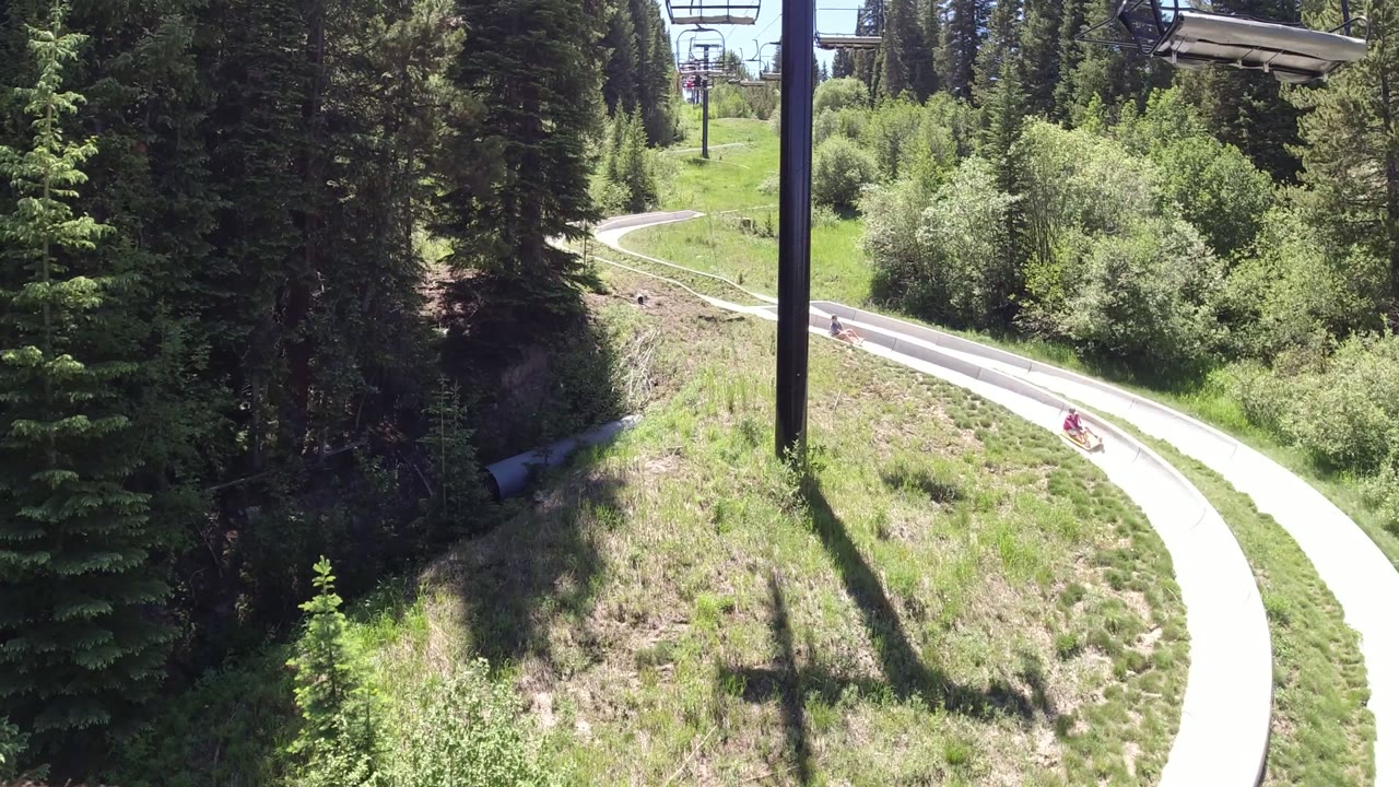 A small rider appears on the upper bend of the go-kart track in Winter Park, Colorado, surrounded by green grass, dense trees, and overhead chairlift hardware.