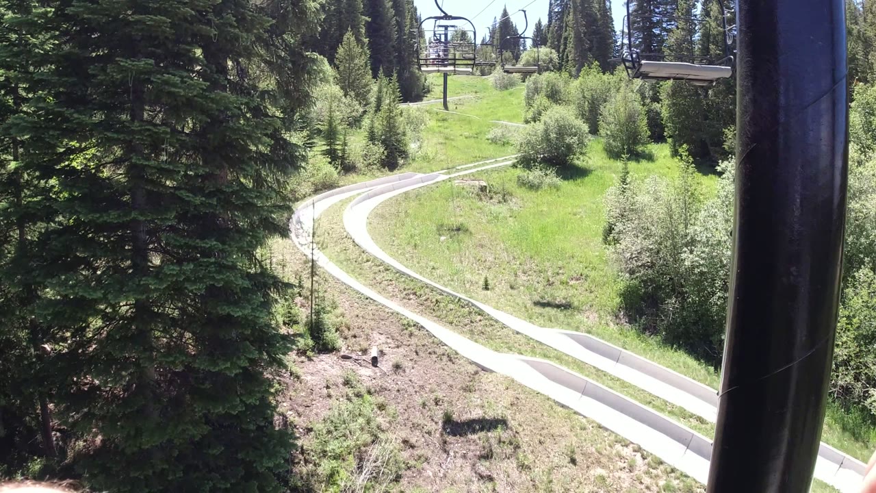 Elevated view in Winter Park, Colorado showing a pale curving track through bright grass, evergreen trees, and chairlift seats overhead with no visible people.