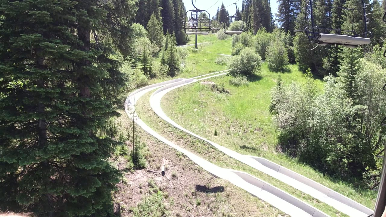 Winter Park, Colorado frame with a dark lift support in the foreground, empty chairlift seats overhead, and a pale track curving between dense green trees with no visible people.