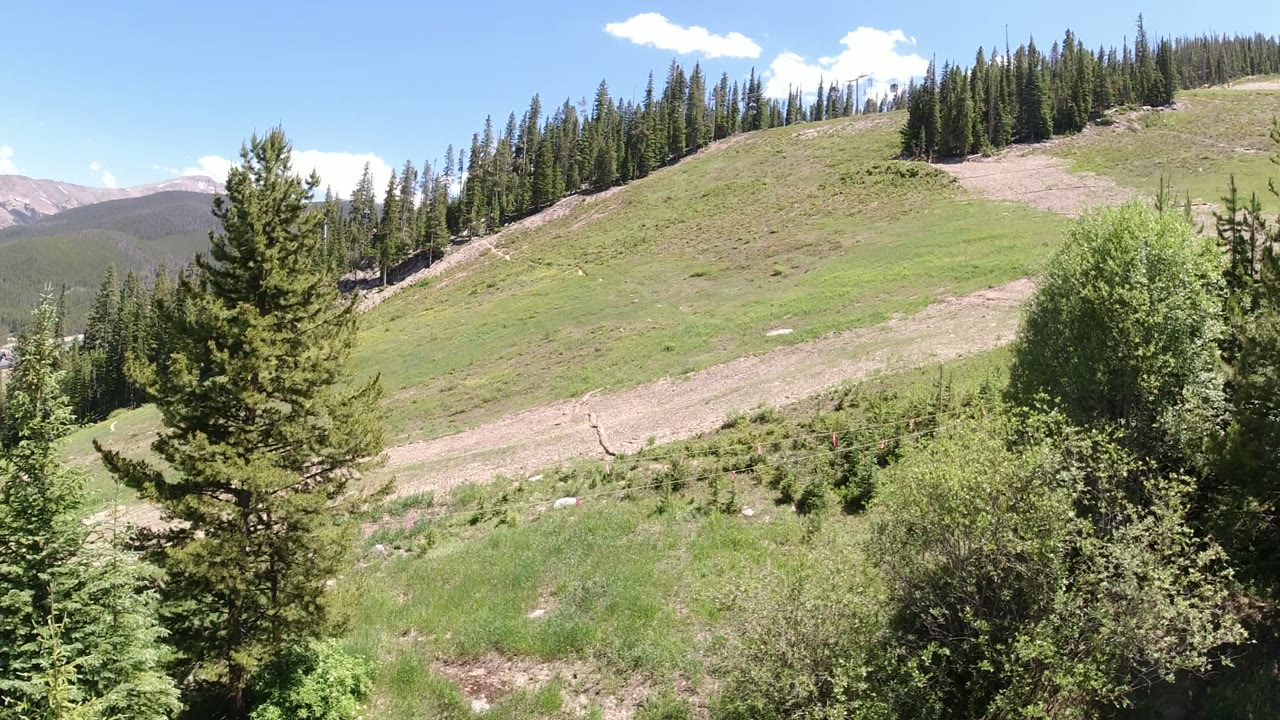 Opening frame of a Winter Park, Colorado mountain slope with foreground evergreens, a grassy hillside, distant ridges at left, blue sky, and no visible people.