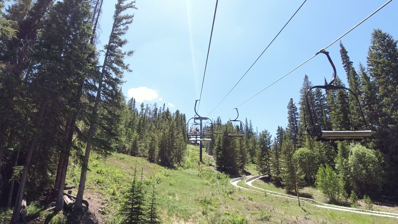 Empty chairlift seats hang closer to center over a grassy Winter Park, Colorado slope, with dense evergreens, a pale winding road, blue sky, and no visible people.