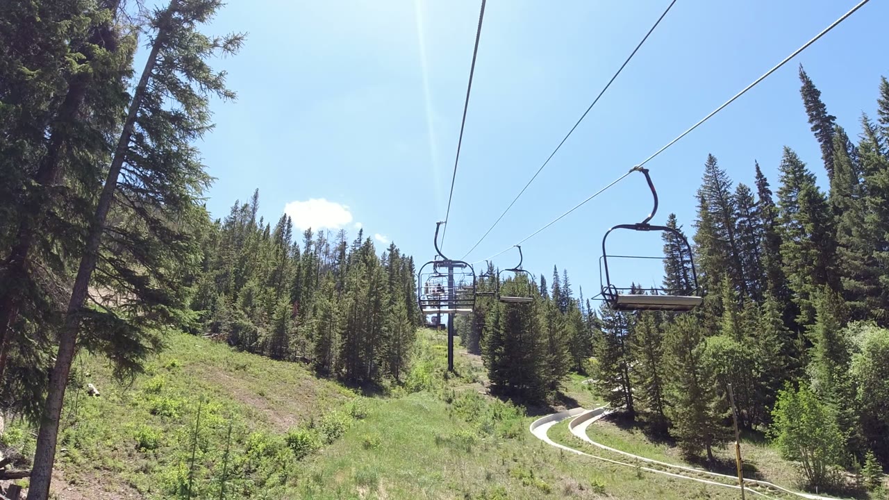 Open chairlift seats run along cables above a green forested slope in Winter Park, Colorado, with a curving path below and no visible people.