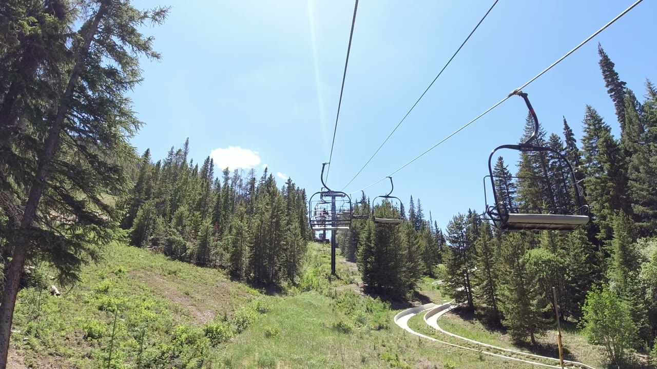 An empty chairlift seat hangs closer to camera on overhead cables in Winter Park, Colorado, with tall evergreen trees and bright sky behind it.