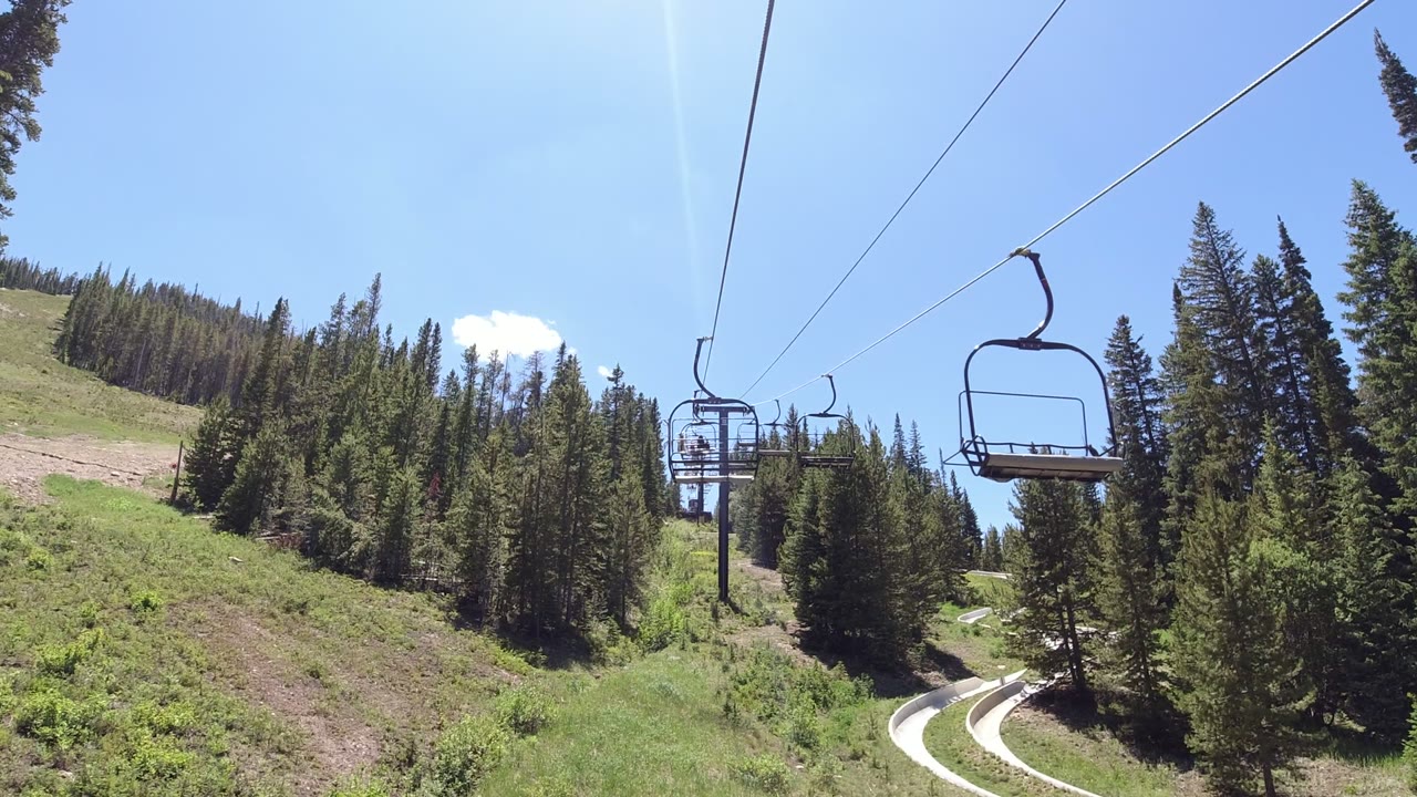 Empty ski lift chairs over a green forested mountain slope.