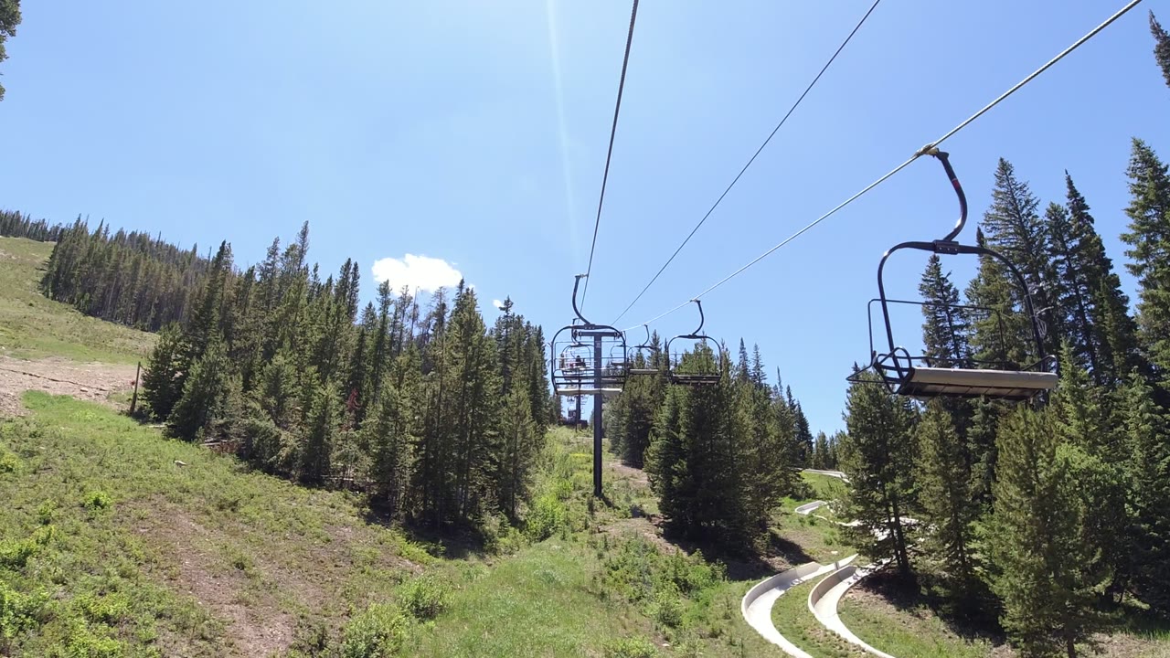 Winter Park, Colorado lift view with the overhead cable leading toward empty chairs, a grassy slope at left, tall pine trees at right, and no visible people.