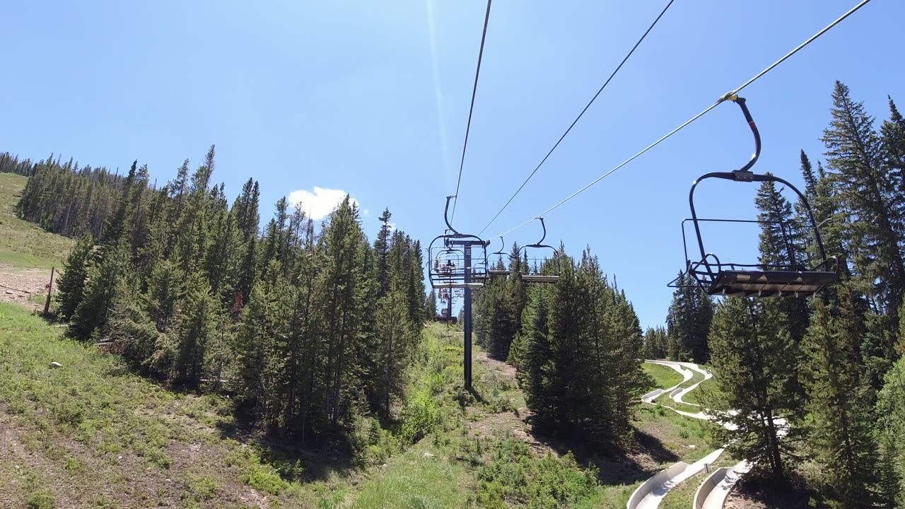 An empty chair passes near the upper center of the frame over conifer trees and hillside terrain in Winter Park, Colorado, with no visible people.