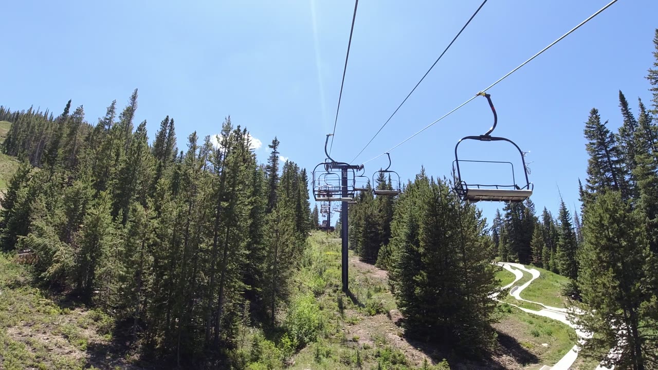 Opening frame of a Winter Park, Colorado chairlift view with cables overhead, empty seats ahead, green pine forest below, a winding pale trail, and no visible people.
