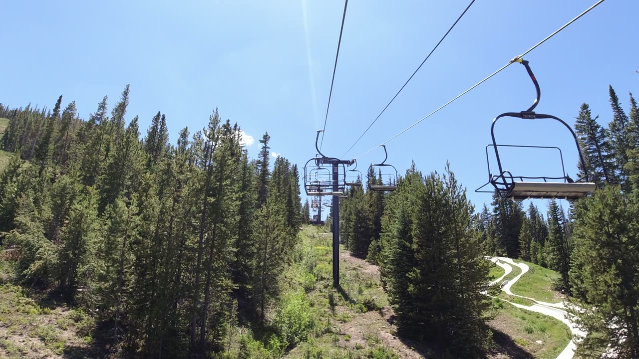 Second frame in Winter Park, Colorado showing an empty chairlift seat near the upper frame, lift cables, dense evergreens, bright sky, and no visible people.