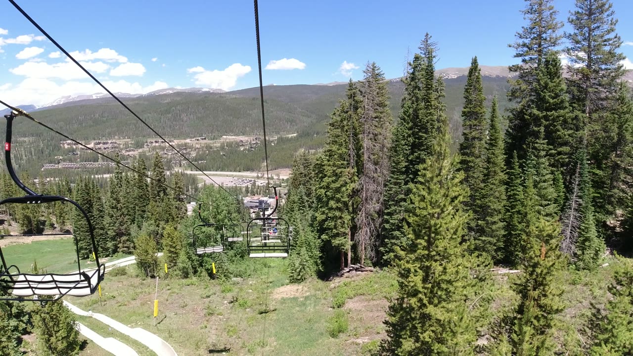 An empty black chairlift seat hangs near the center of the Winter Park, Colorado view, with cables overhead, green trees below, and mountain ridges in the distance.