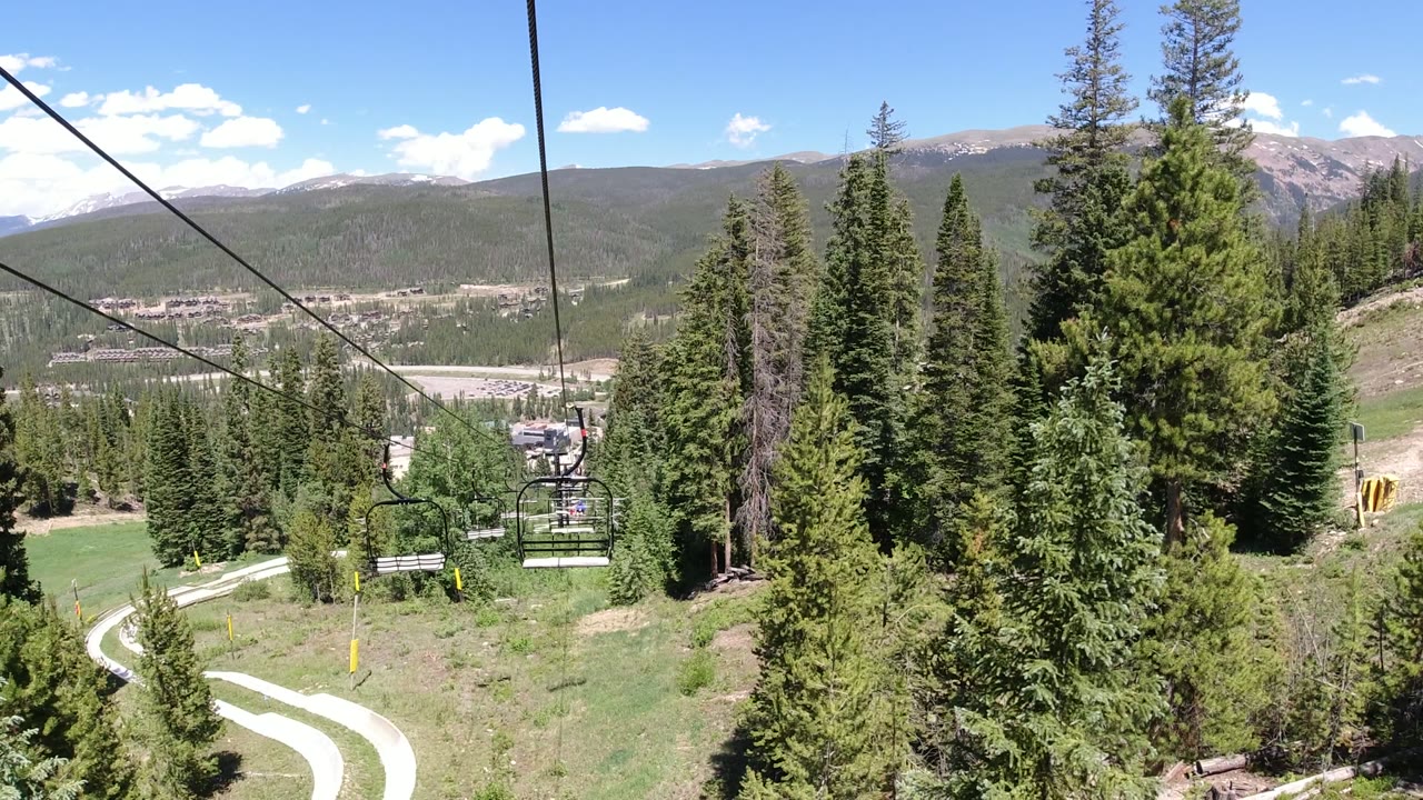 Opening view in Winter Park, Colorado, with lift cables stretching over conifer trees, an empty chair at lower left, distant ridges, blue sky, and no visible people.
