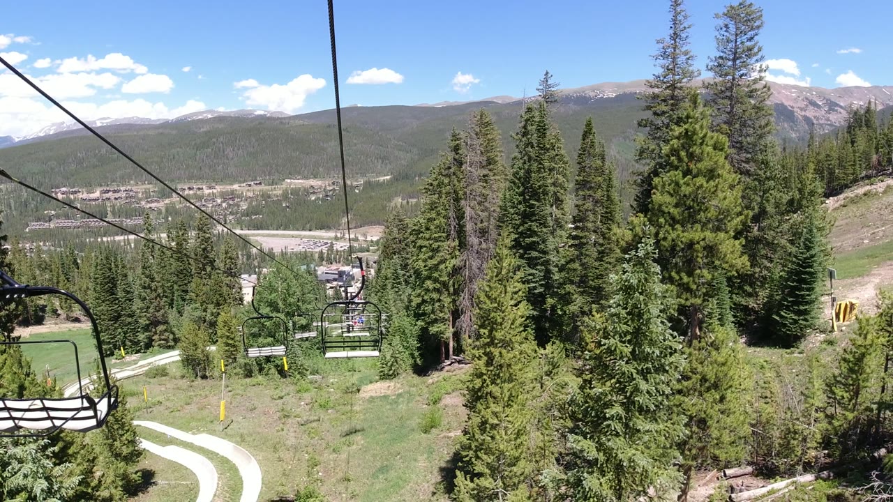 Second frame shows an empty chairlift seat nearer the center-right, black cables overhead, dense evergreens below, a pale curved track, and no visible people.
