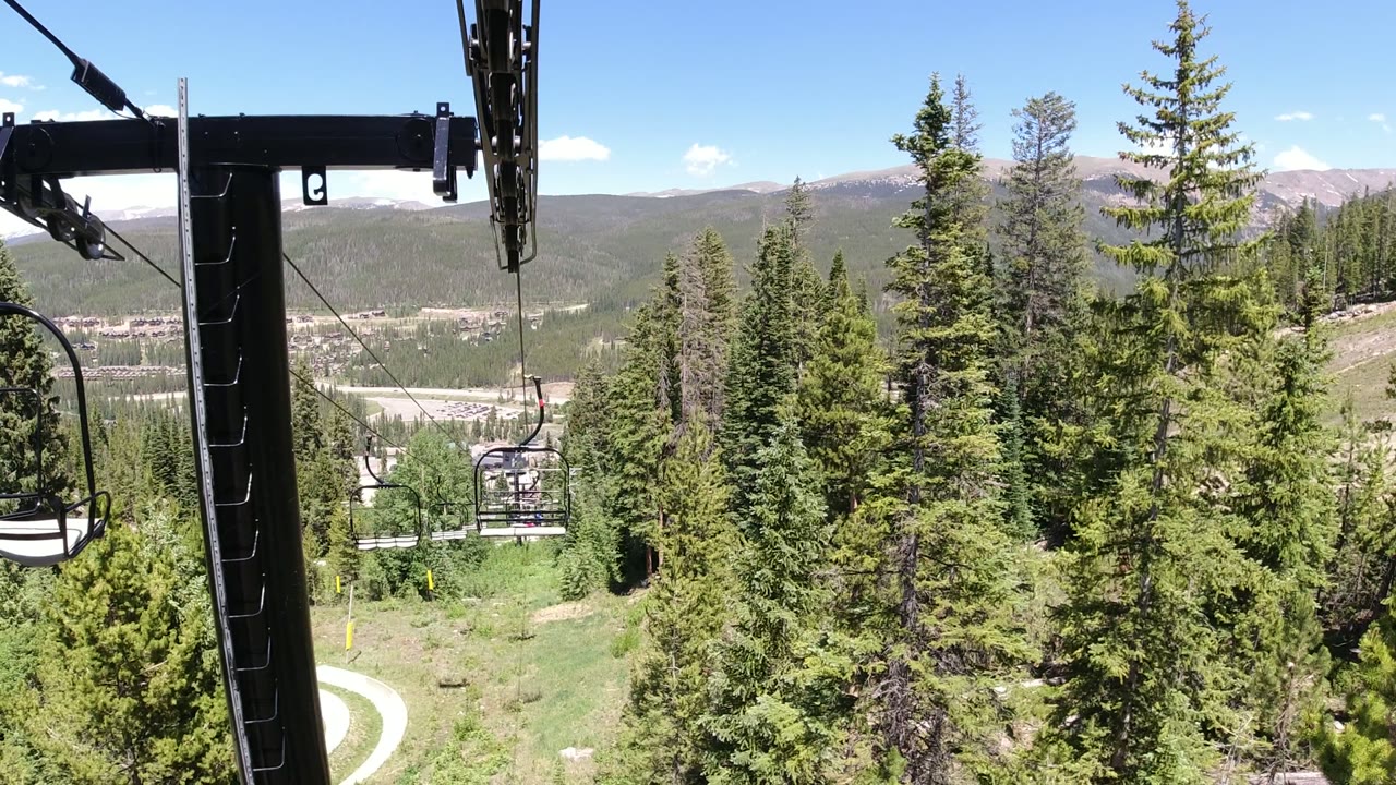 Winter Park, Colorado lift line with a black support arm near the center, chairlift seats suspended over evergreens, pale mountain ridges in the distance, and no visible foreground people.
