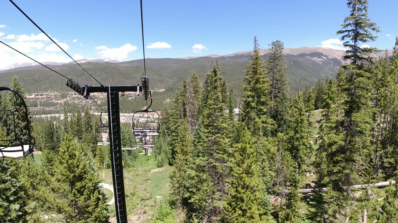 An empty-looking chairlift seat hangs beside a black lift tower above dense trees in Winter Park, Colorado, with a valley and mountain ridge beyond.