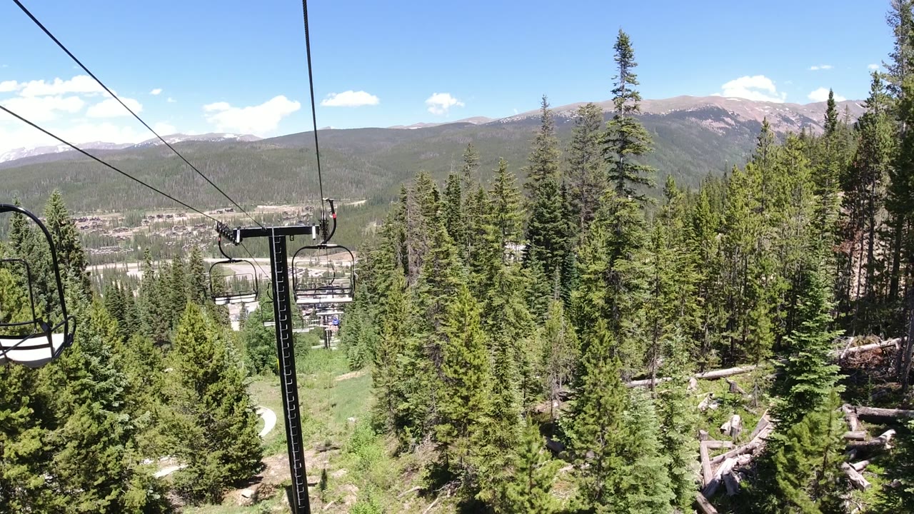 A nearby empty chair hangs from the cable above dense green trees in Winter Park, Colorado, with a pale valley and mountain slopes beyond.