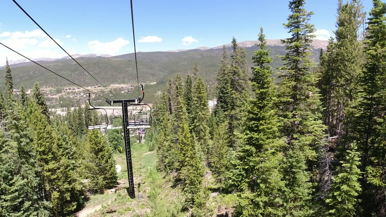 A Winter Park, Colorado chairlift cable crosses above dense green pine trees, with an empty lift chair partly visible and no visible people in this frame.