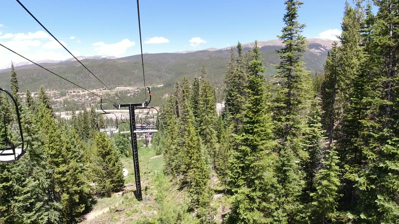 An empty chairlift seat hangs closer to the center of the Winter Park, Colorado frame, with tall pines below and a hazy mountain valley beyond, with no visible people.