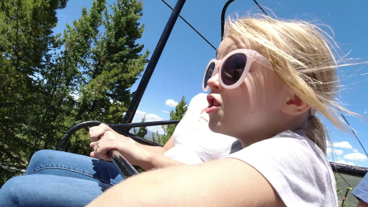 At the start of the Winter Park, Colorado clip, a blonde girl in pink sunglasses sits close to the camera on a ski lift, with trees, blue sky, and black lift bars behind her.