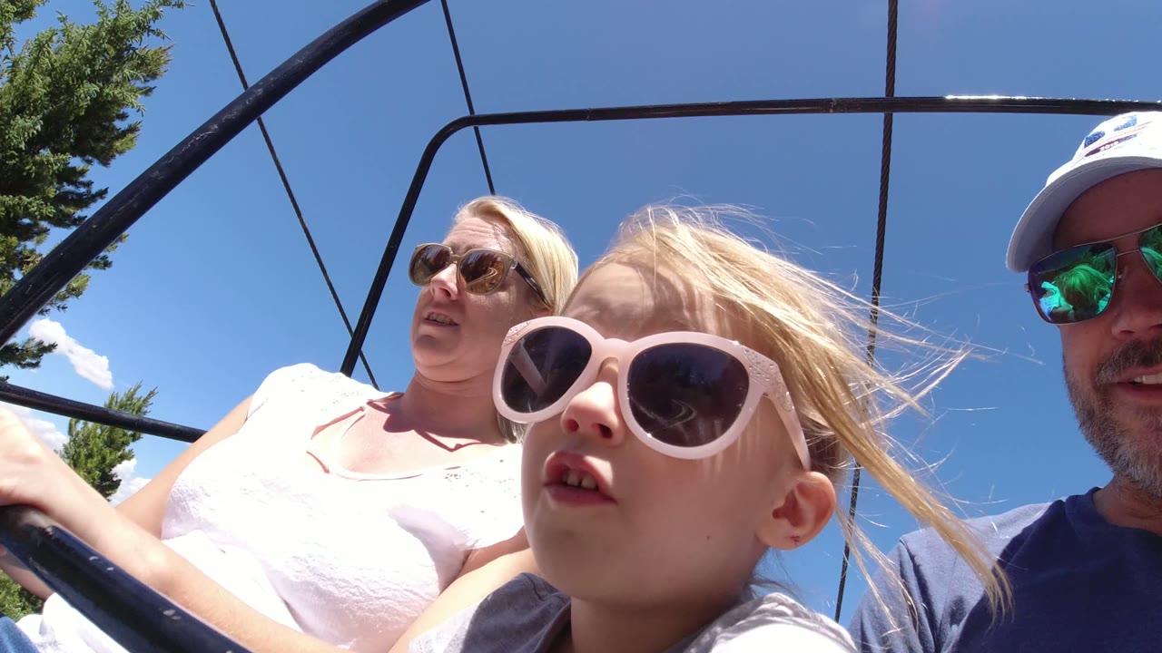 One second into the Winter Park, Colorado clip, the woman and girl sit on the ski lift while part of the man enters the right edge, all under clear blue sky and black overhead bars.