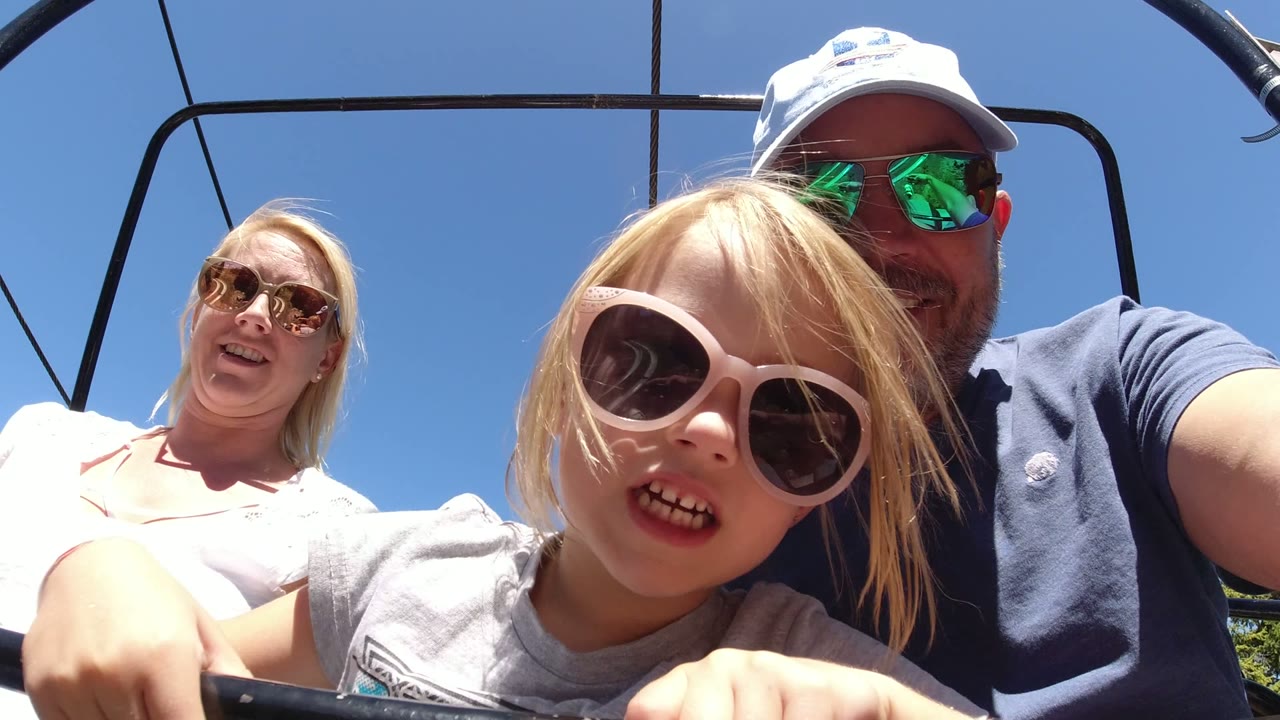 The girl remains centered in the foreground as both parents are more visible on the open chairlift in Winter Park, Colorado, with lift bars silhouetted above.