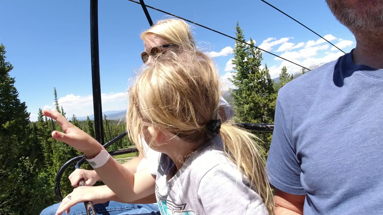 The child turns and raises a hand beside the chairlift rail while adults sit nearby over the pine forest in Winter Park, Colorado.