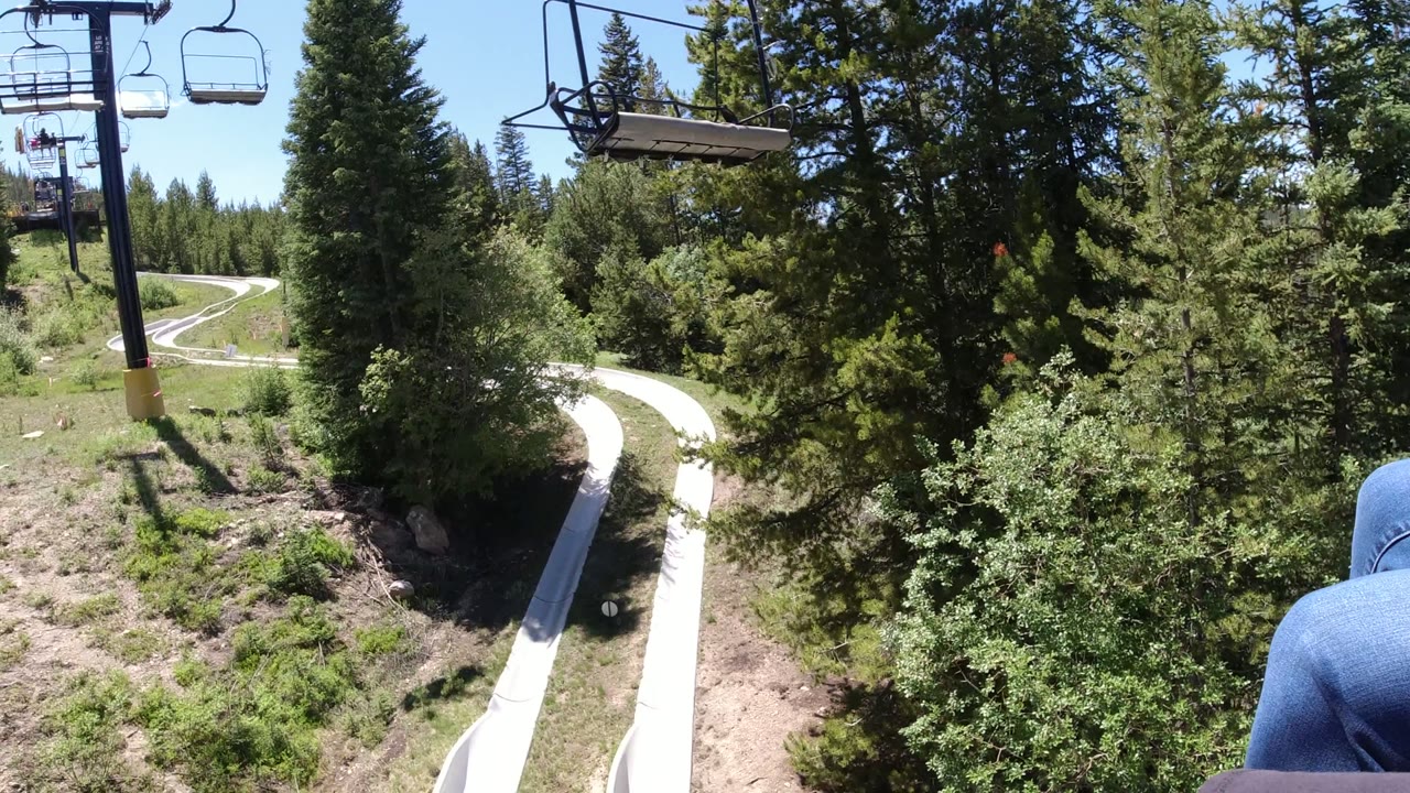 Chairlift view over a curving alpine slide track and evergreen trees.
