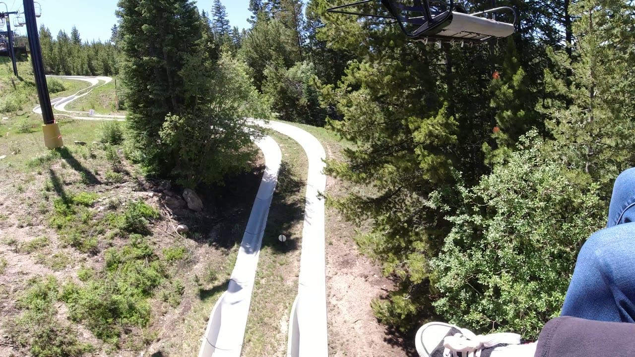 Winter Park, Colorado chairlift view with the white alpine slide track bending below, trees filling the frame, and part of a rider's blue-jean leg at the right edge.