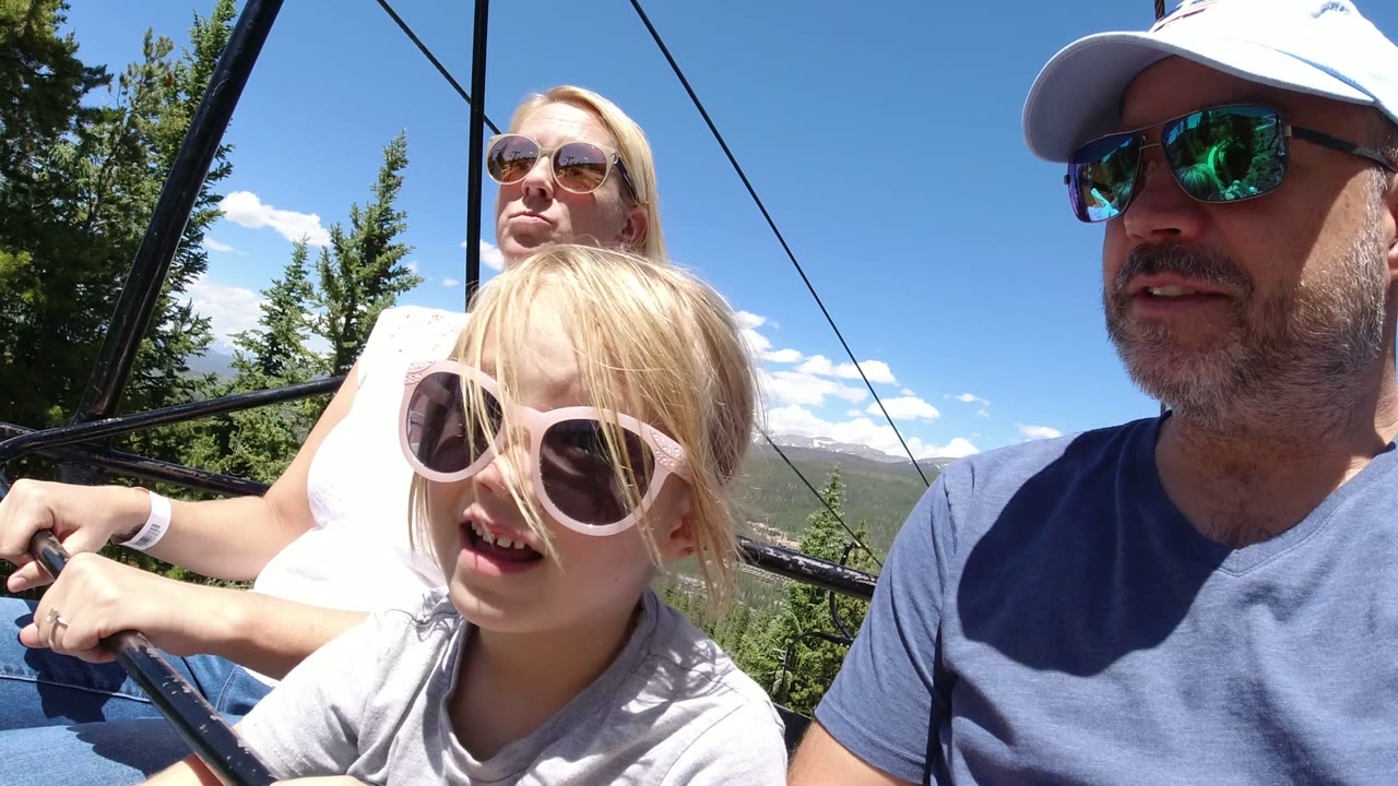 Family riding an open chairlift with evergreen trees behind them.
