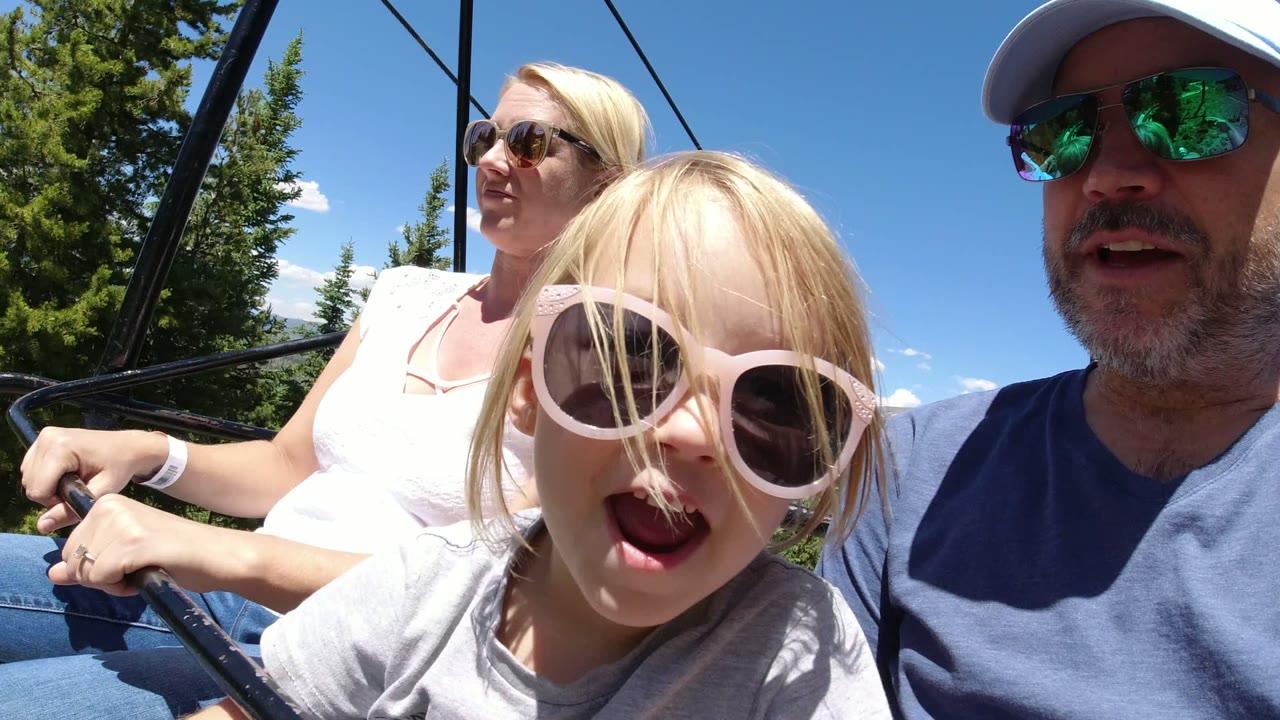The Winter Park chairlift ride continues with the child leaning nearer the lens, both adults partly visible, and sunlit conifers filling the background.