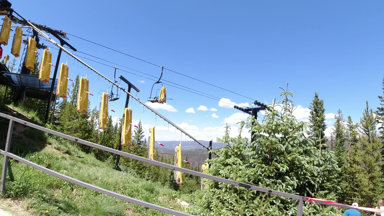 Yellow padded lift covers hang from cables above a grassy slope in Winter Park, Colorado, with evergreen trees, railings, and distant mountains under a blue sky, no visible people.