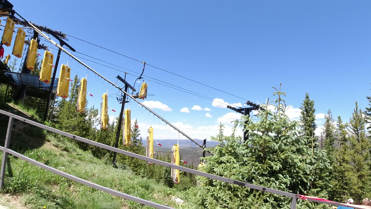 A similar Winter Park, Colorado hillside view shows yellow lift covers, overhead cable lines, a metal railing, evergreen trees, and distant mountain ridges, no visible people.