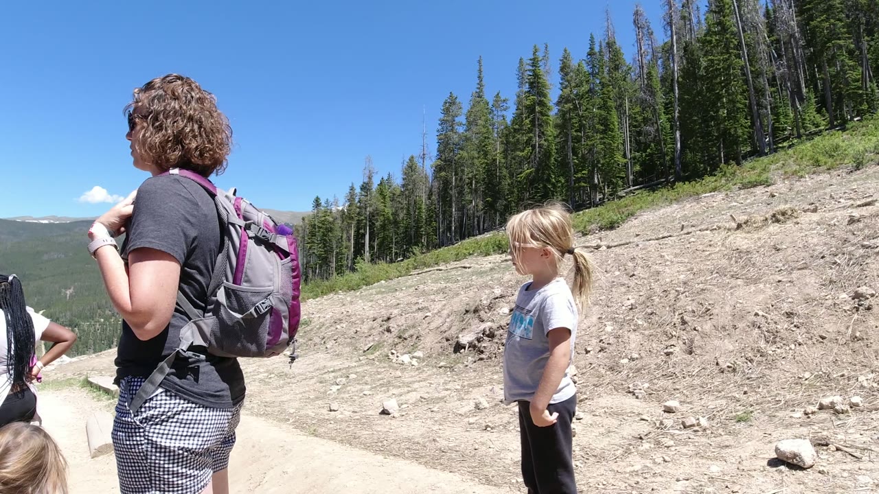 An adult with a gray and purple backpack stands near a young girl on a dirt road in Winter Park, Colorado, with trees and distant mountain terrain behind them.