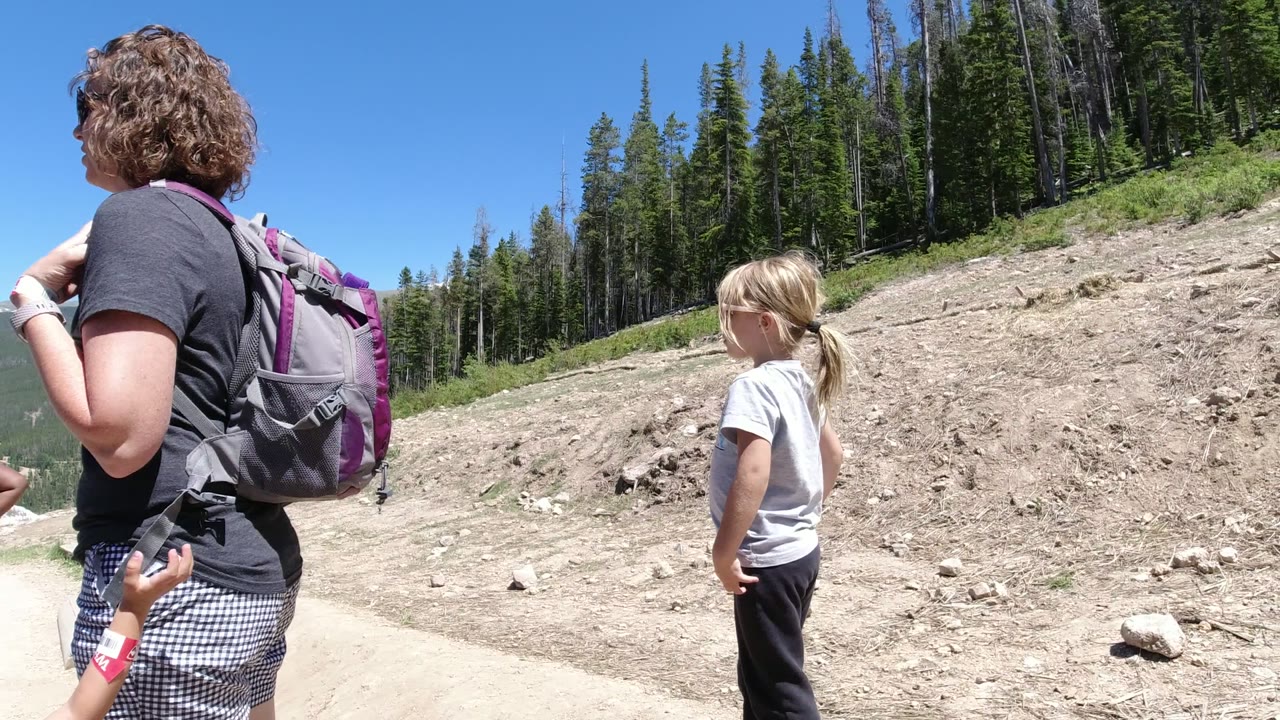 The young girl stands near the center of the dirt road scene while a backpacked adult is partly visible in Winter Park, Colorado, with evergreen forest rising on the slope.