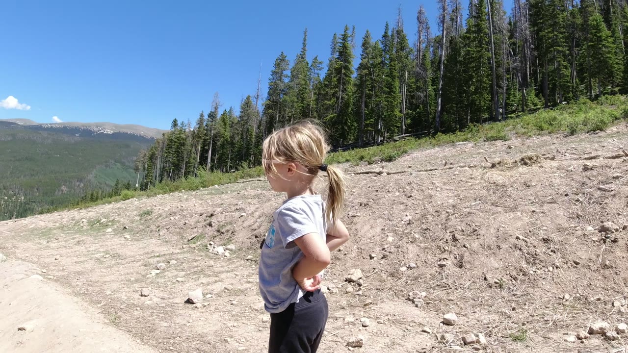 Young girl standing on a dirt mountain trail beside evergreen forest.