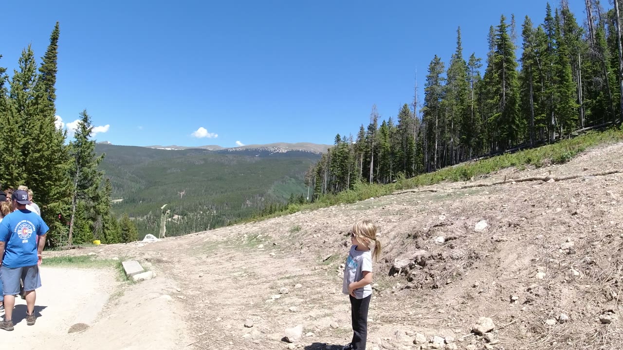 At Winter Park, Colorado, a girl in the foreground and a man in a blue shirt stand beside a gravel path, with a rocky slope, evergreen trees, and distant mountains behind them.