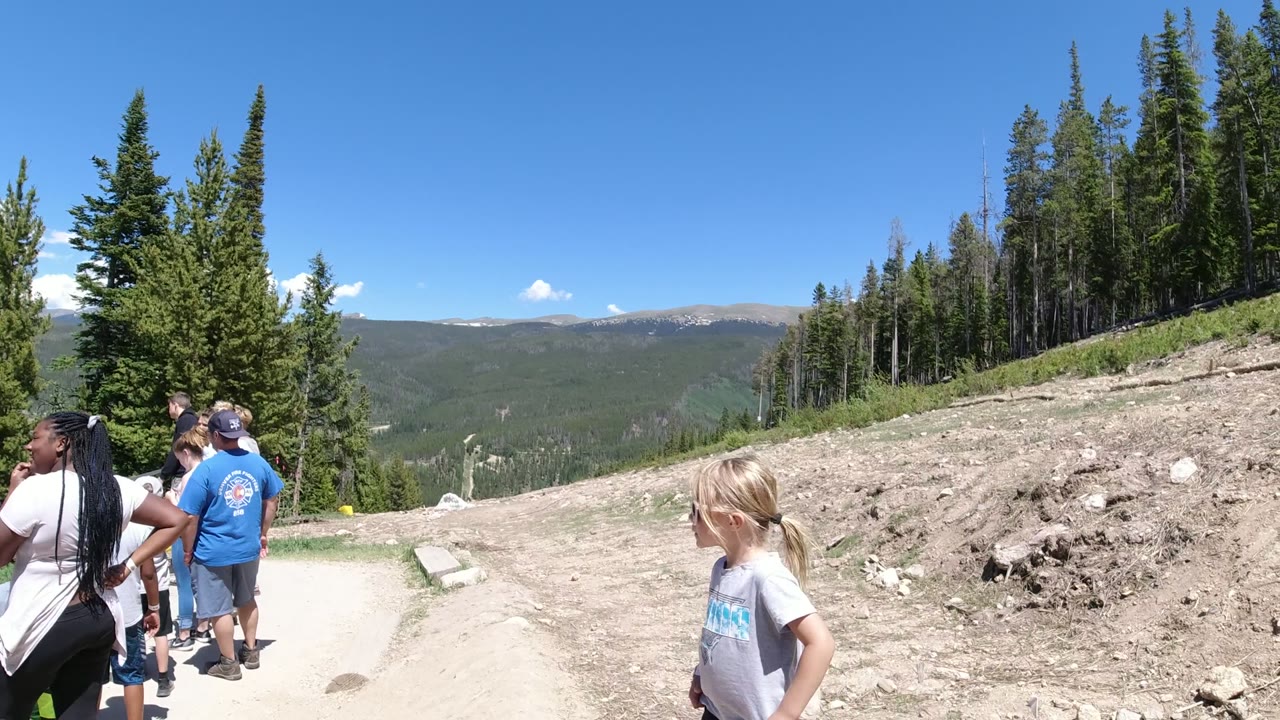 People standing on a dirt mountain trail with evergreen trees and distant ridges.