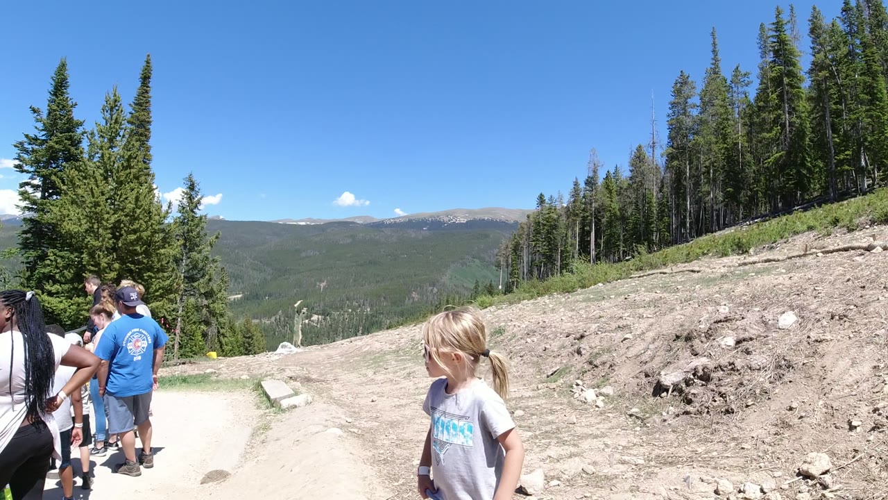 At 0:01 in Winter Park, Colorado, adults gather near the left side of the trail while the child remains on the right foreground, with a sloped dirt bank, conifers, and distant mountains in bright sun.