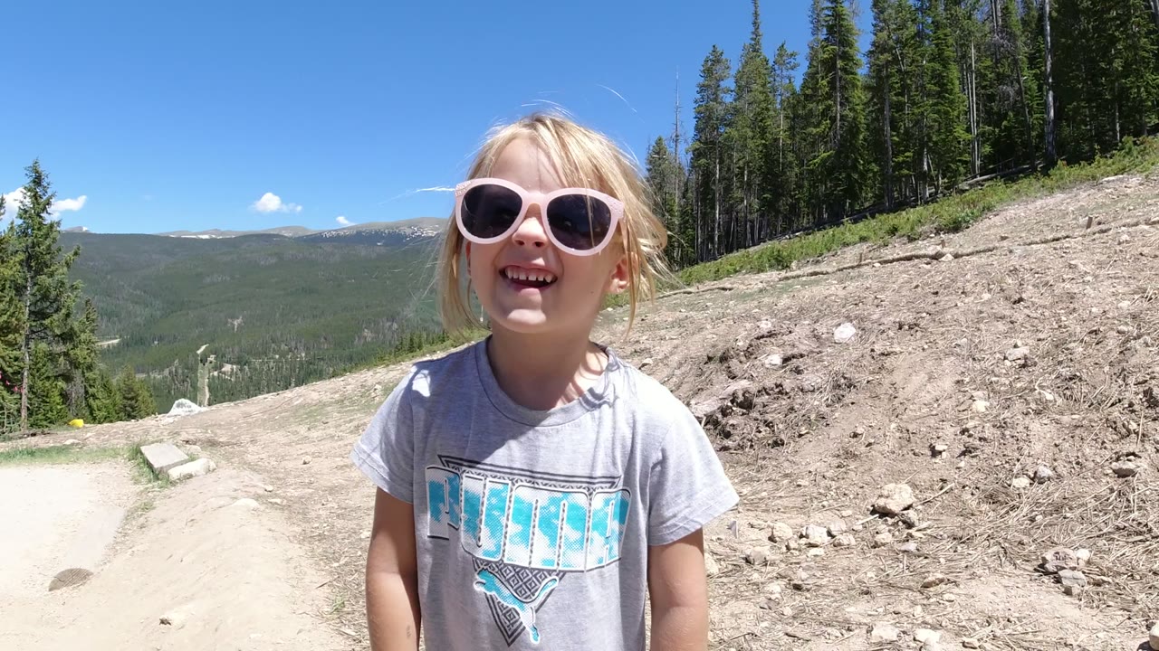 A blond child in pink sunglasses stands near the camera on a dirt road in Winter Park, Colorado, with evergreen trees, rocky ground, distant hills, and blue sky behind her.
