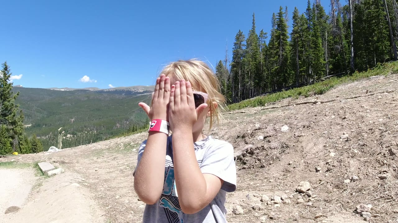 The child raises both hands over her pink sunglasses in Winter Park, Colorado, while standing beside a dirt road with forested slopes and open mountain views in the background.