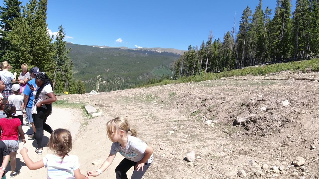 Children and adults gather on a dirt path with forested hills behind them.