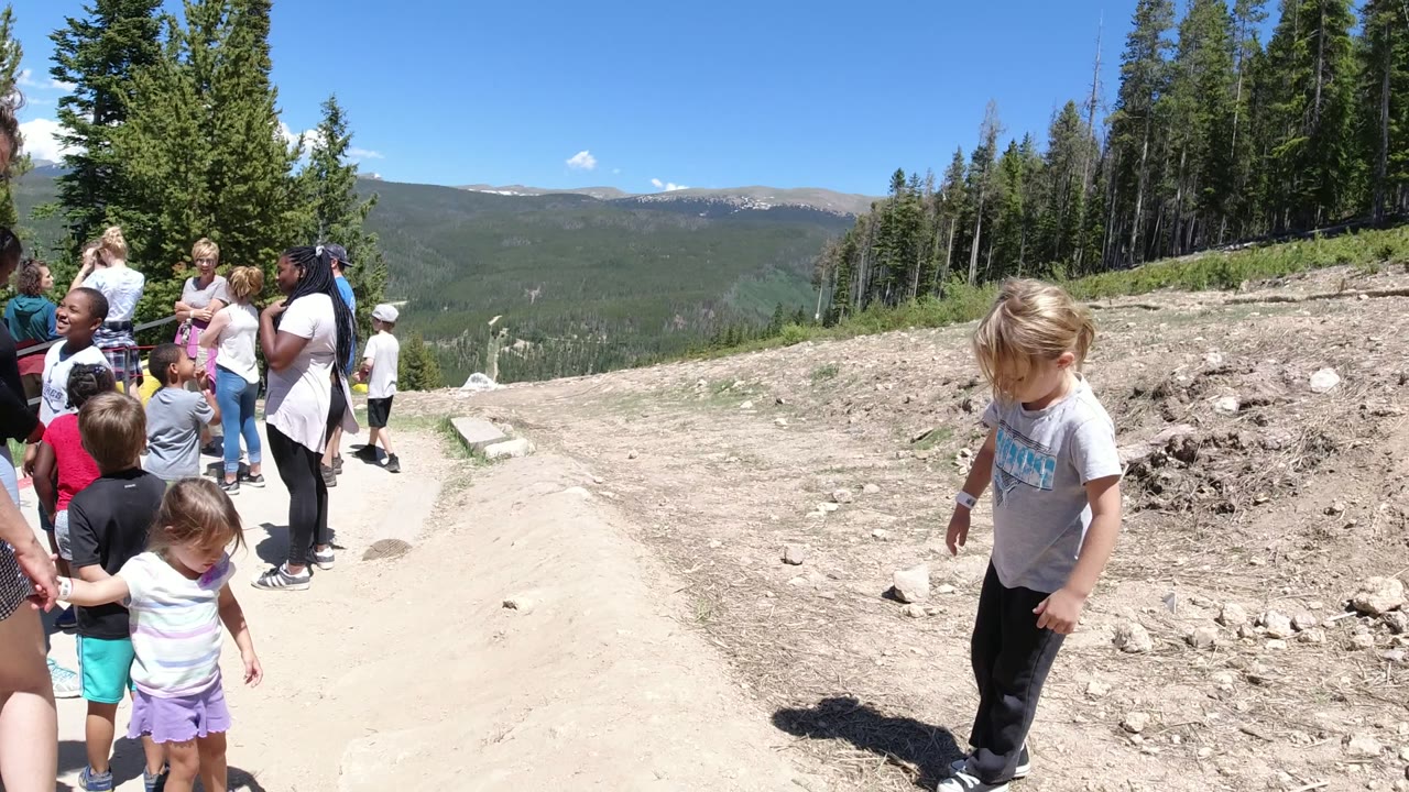 At the start of the clip in Winter Park, Colorado, several children and adults stand and walk along a bright dirt mountain path with evergreen trees and distant ridges behind them.