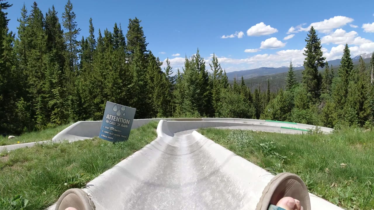 The Winter Park, Colorado slide path runs forward between grass and trees, with a readable rain warning sign beside the concrete and a sandaled foot at the lower edge.